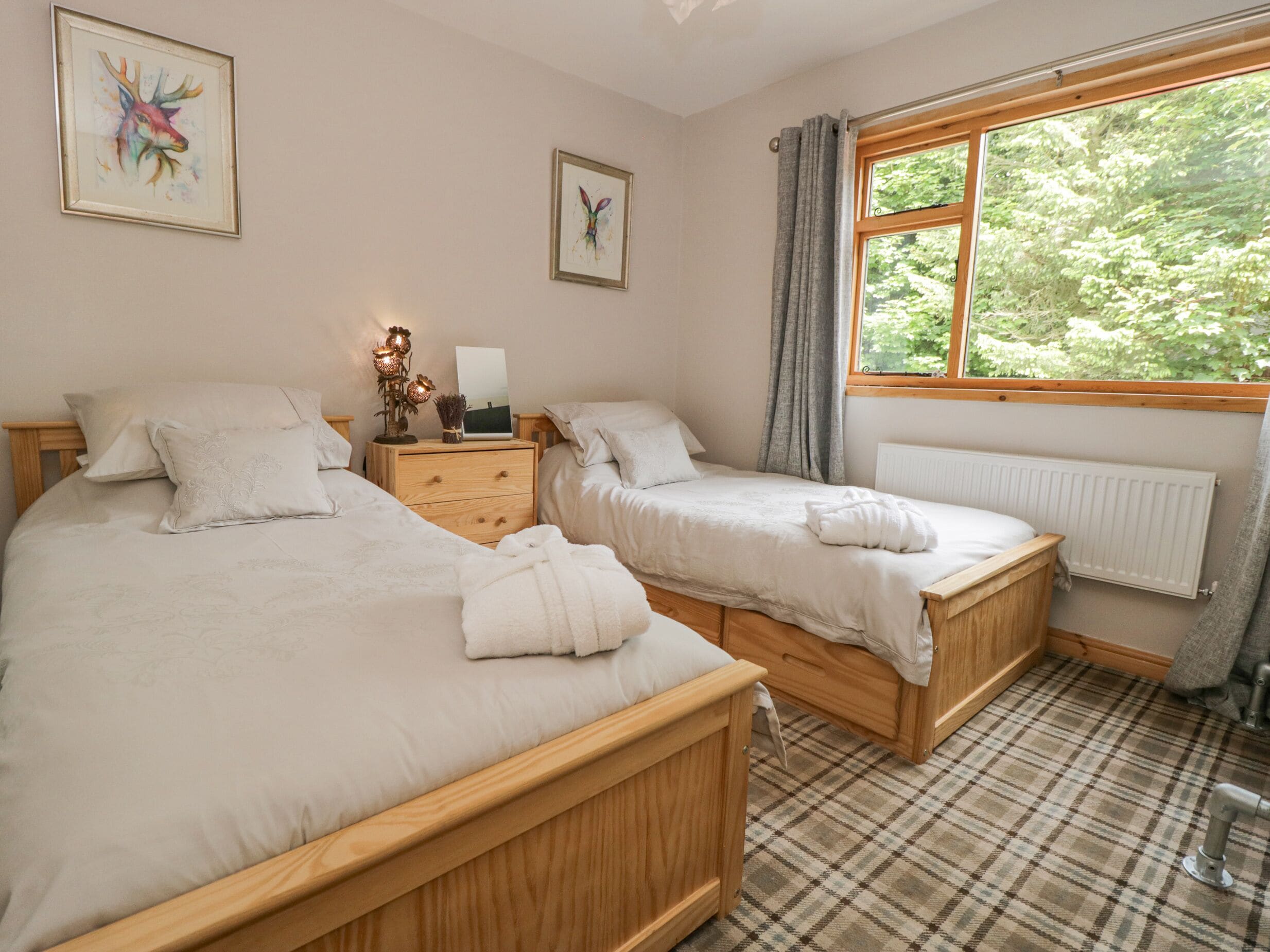 A bedroom with two wooden beds, a nightstand with a lamp, and a window with a view of greenery.