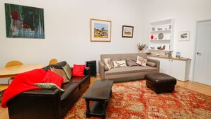 A living room with a sofa, a coffee table, and a patterned rug. There is a painting on the wall and a small cabinet with decorative items.