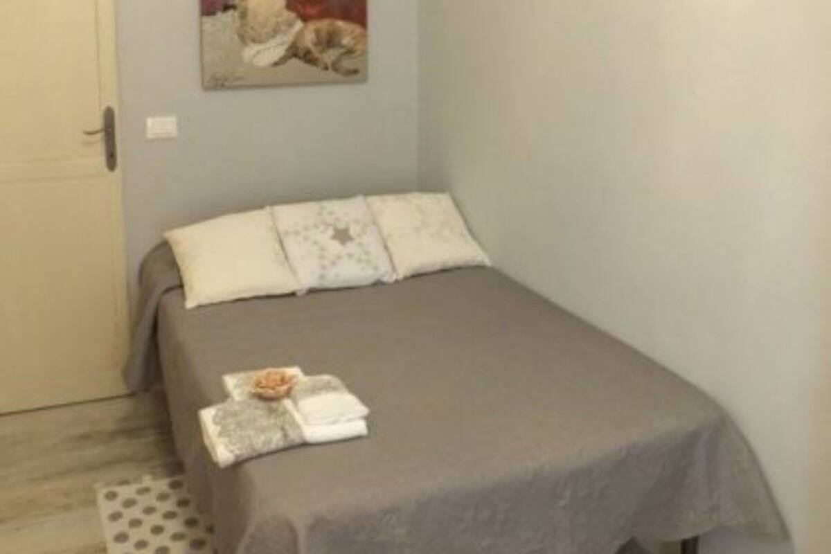 Un petit lit bien fait, avec une couverture grise et des oreillers blancs, une petite table sur laquelle est posée une serviette pliée et un livre, et une image d’un chat accrochée au mur.