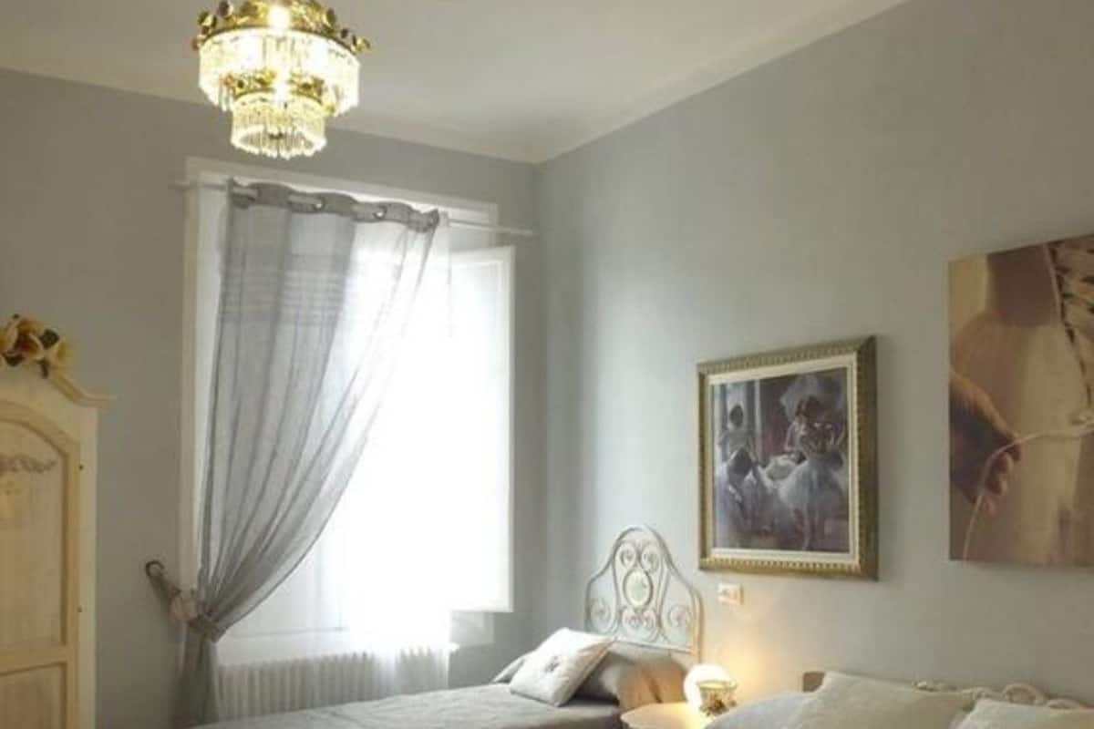Une chambre avec deux lits, un lustre, une fenêtre avec des rideaux, un radiateur et un tableau encadré au mur.