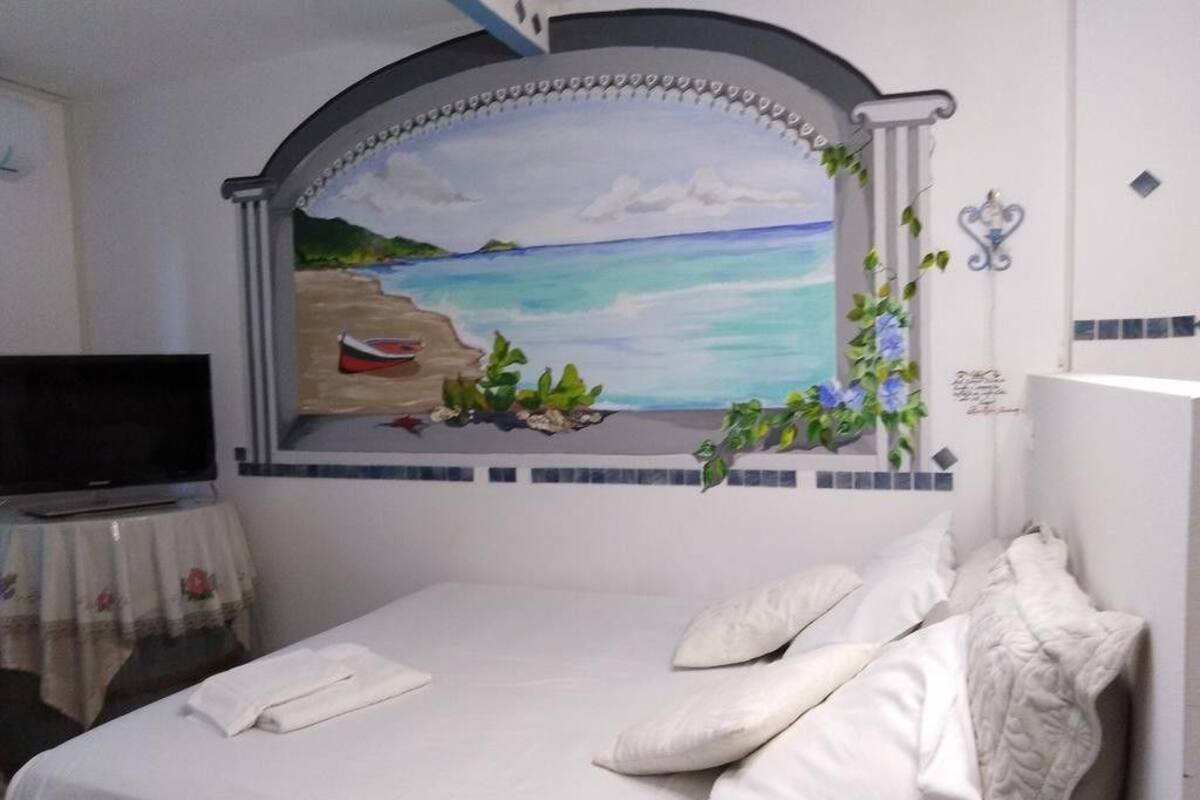 Une chambre à coucher avec un lit, une télévision, un tableau représentant une scène de plage et une petite table recouverte d’une nappe à motifs floraux.
