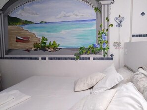 Un dormitorio con cama, ropa de cama blanca, un cuadro de una playa y una pared decorativa con una columna y plantas.
