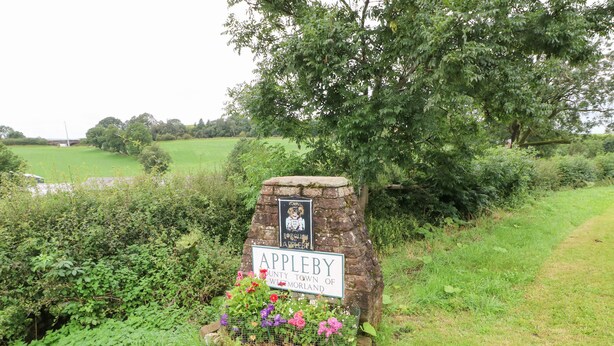 Un letrero de piedra con la inscripción 'Appleby' y 'Ciudad del Condado de Westmorland', rodeado de follaje y flores.