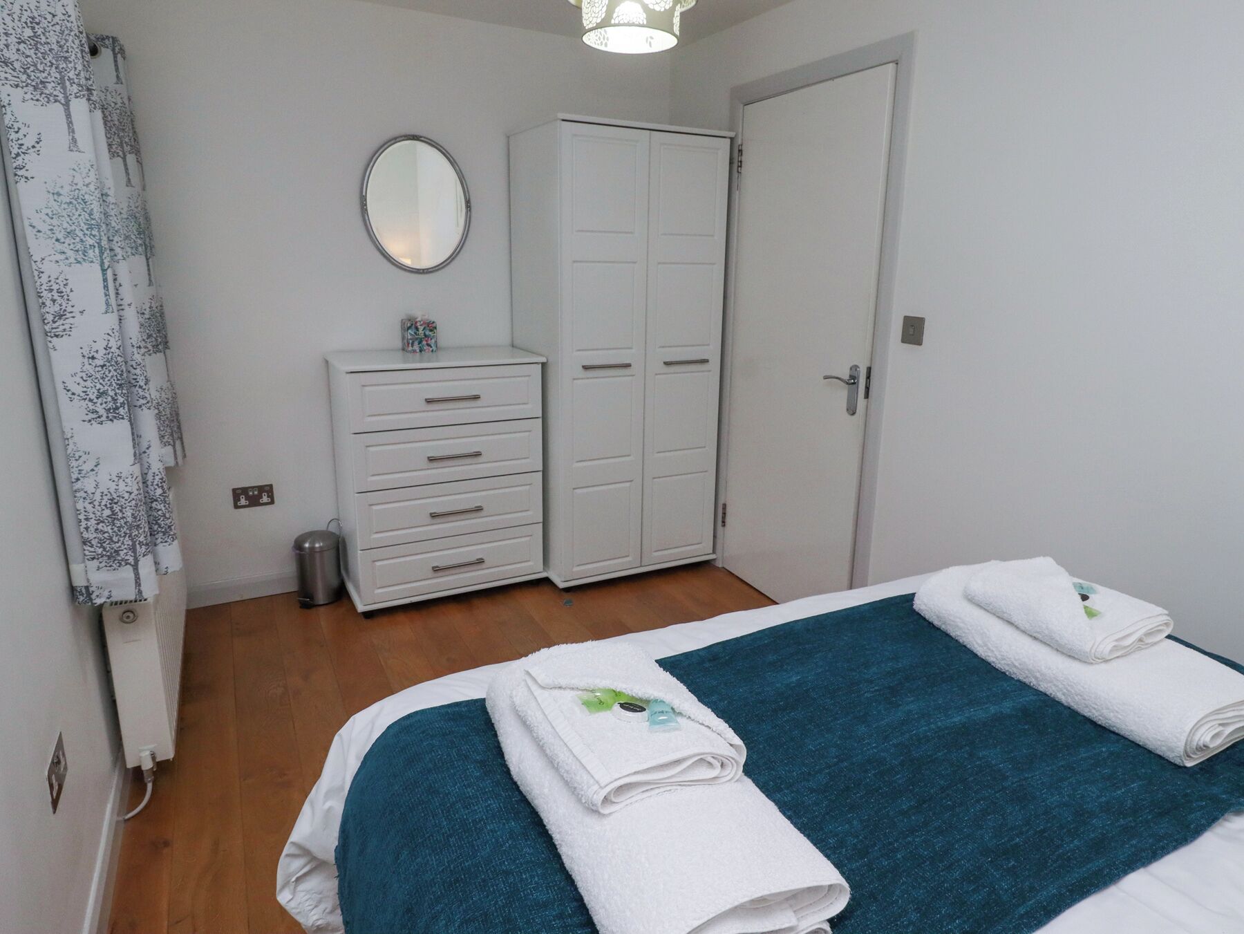 A bedroom with a bed, two white towels, a blue blanket, a white wardrobe, a mirror, and a radiator.