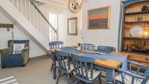 A dining area with a wooden table and blue chairs, a staircase, a bookshelf with nautical decor, and a framed painting of a sunset. - Marsh Villa (Wigton)