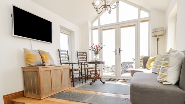 A modern living room with a grey sofa, wooden flooring, a TV mounted on the wall, and a dining area with wooden chairs and a table. - Marsh Villa (Wigton)