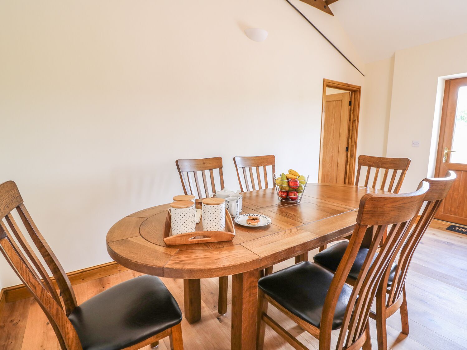A wooden dining table with six chairs, a fruit basket, and a tray with mugs and a plate of food.