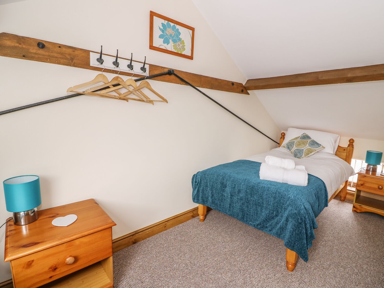 Un dormitorio con cama, mesitas de noche de madera, perchero y un cuadro en la pared, además de una lámpara azul.
