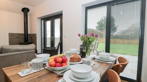 Un comedor con una mesa de madera dispuesta para cuatro personas, rodeada de sillas. Se ve una ventana grande con persianas, y una chimenea al fondo.