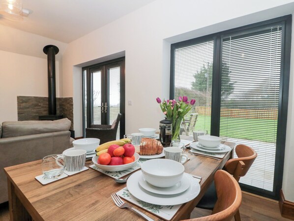 A dining area with a wooden table set for four, surrounded by chairs. A large window with blinds is visible, and a fireplace is seen in the background. - Fieldside (Filey)