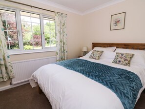 A bedroom with a large bed, a window with curtains, a radiator, a bedside table with a lamp, and a framed picture on the wall. - Pen Y Maes (Builth Wells)