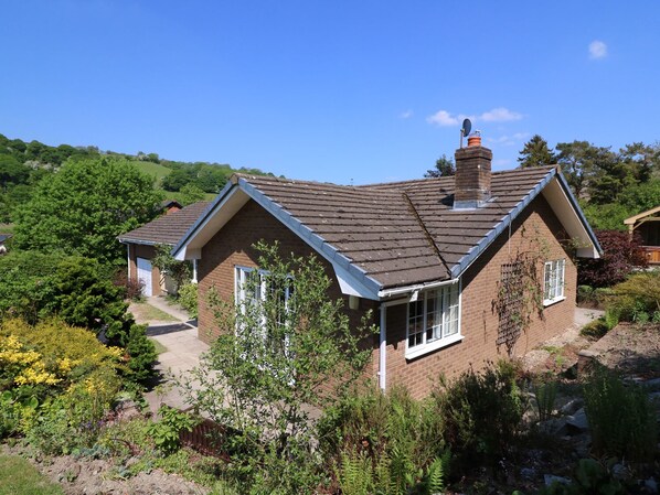 A two-story house with a sloped roof, a brick exterior, and a chimney. There is a garden with various plants and flowers in front of the house. - Pen Y Maes (Builth Wells)