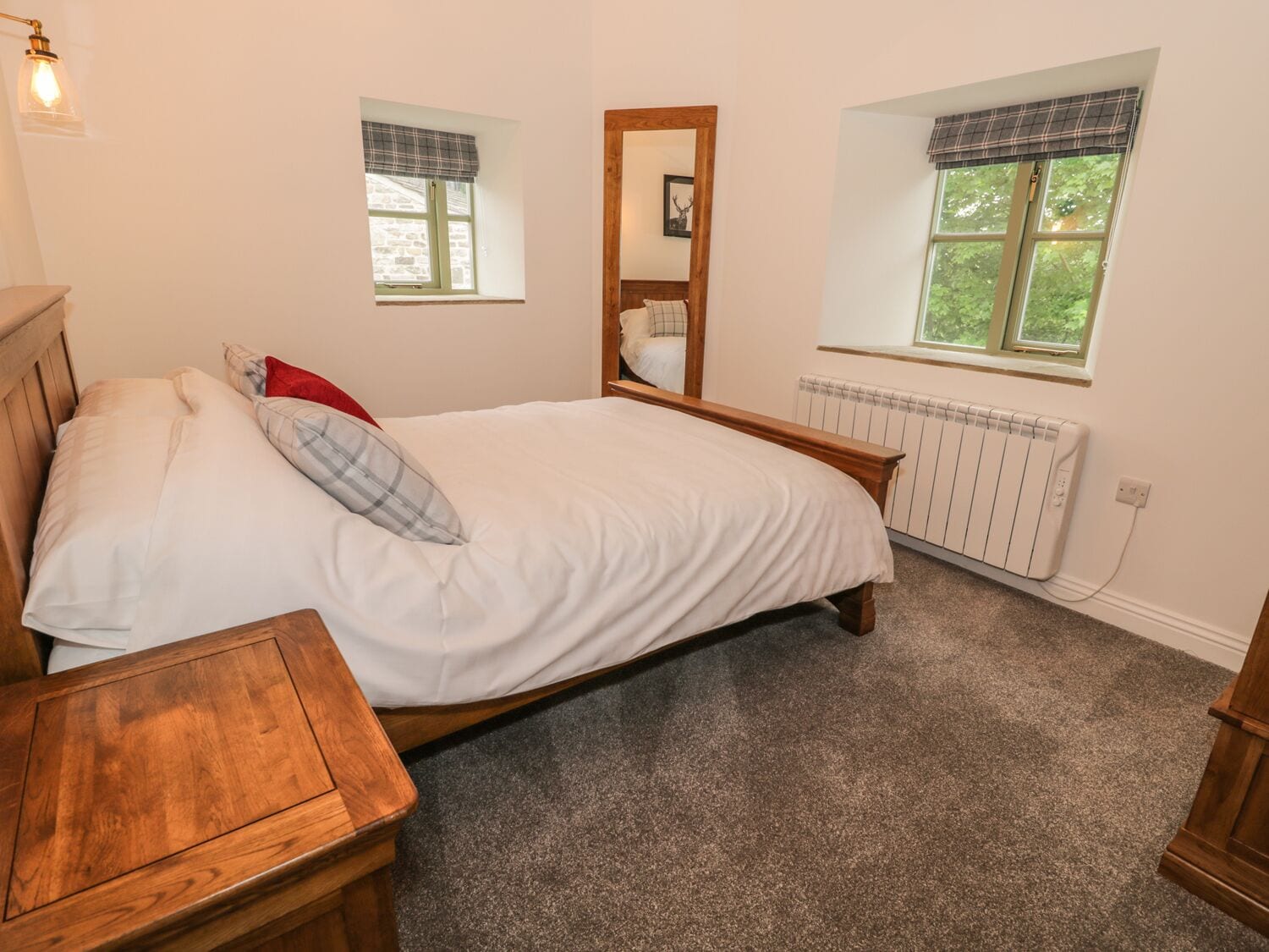 A bedroom with a wooden bed, a nightstand, a mirror, two windows with curtains, and a radiator.