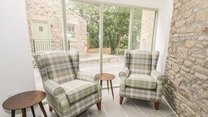 A cozy room with two armchairs and a small table, large windows offering a view of a stone building and greenery outside. - Oak Cottage (Barnard Castle)