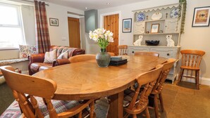 A dining area with a wooden table and chairs, a vase with flowers, and a decorative shelf with plates and a teddy bear. - Trefollwyn Mawr (Pwllheli)