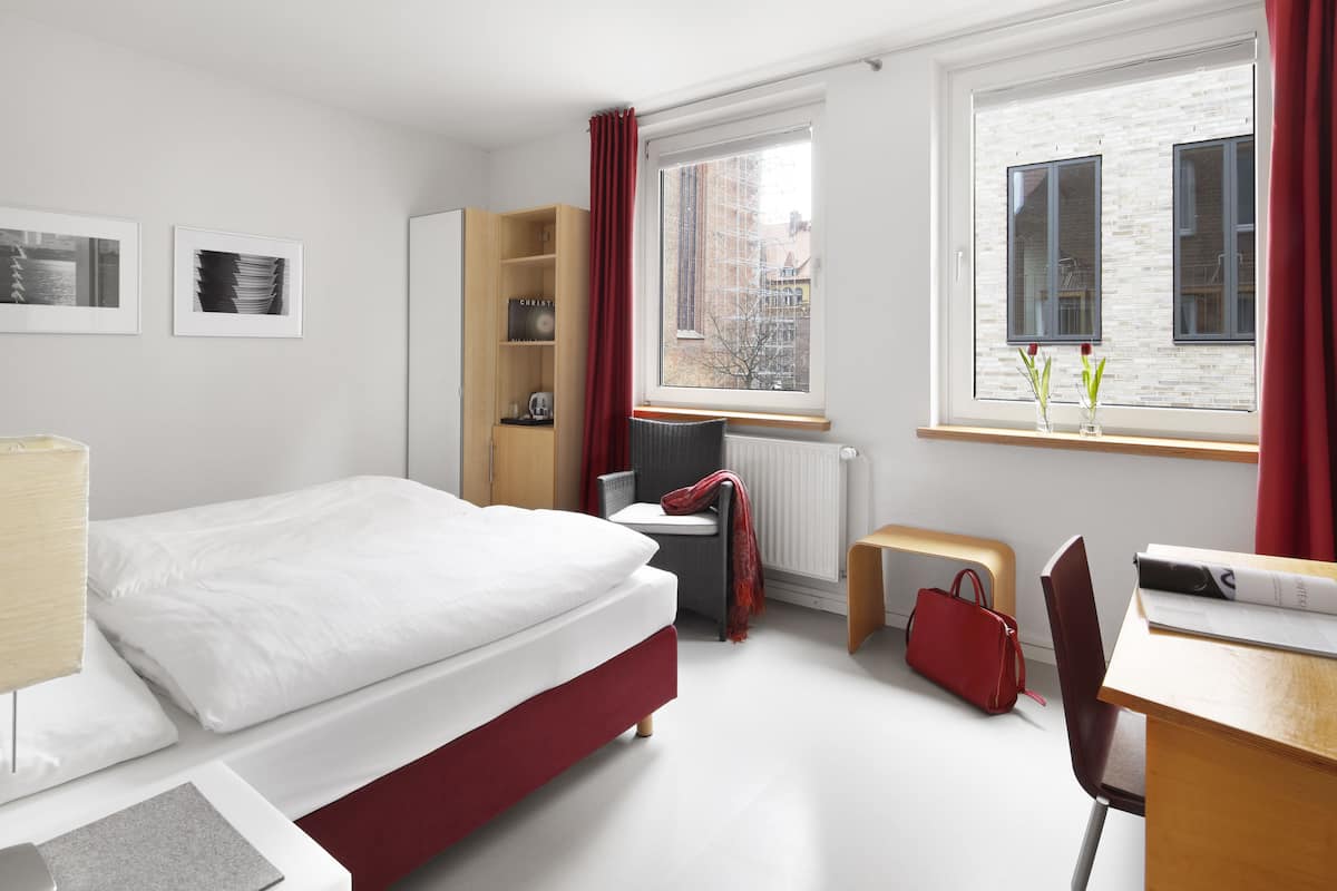 A modern hotel room with a bed, a desk, a chair, a small table, a radiator, a window with curtains, and a bookshelf.