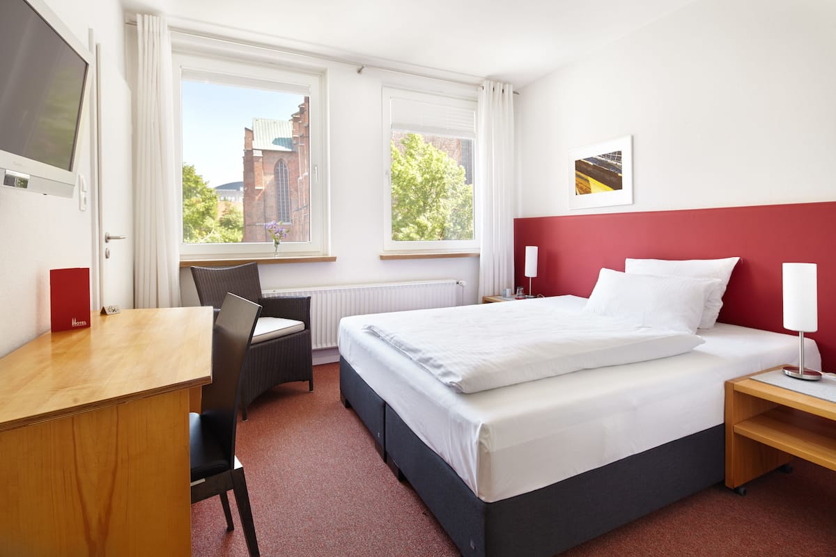 A hotel room with a bed, desk, chair, and two windows offering a view of buildings and trees.