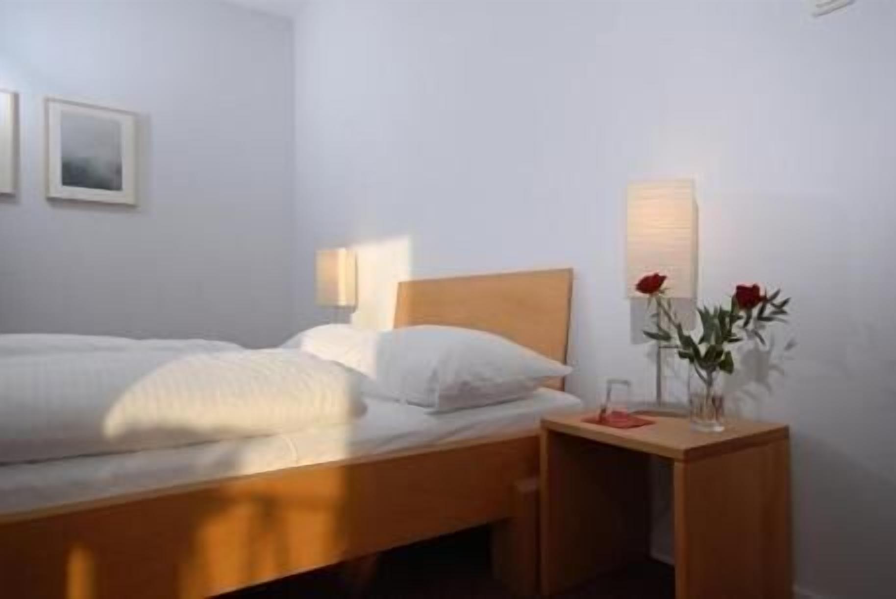 A neatly made bed with white linens, a wooden headboard, a bedside table with a vase of red roses, and a wall-mounted lamp.