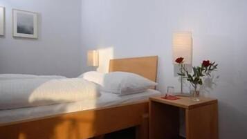 A neatly made bed with white linens, a wooden headboard, a bedside table with a vase of red roses, and a wall-mounted lamp.