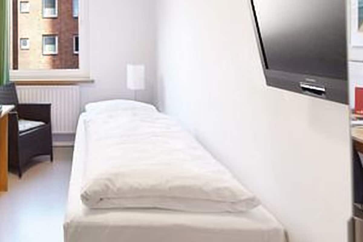 A small, compact hotel room with a single bed, a desk, and a chair. There is a window providing a view of the outside.