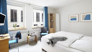 A modern hotel room with a bed, a desk, a chair, a small table with a tray of wine glasses, and a bookshelf. - Hotel an der Marienkirche (Lübeck)