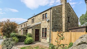 A stone house with a front porch, a wooden fence, and a garden with shrubs and a tree. - Croft Cottage (Skipton)