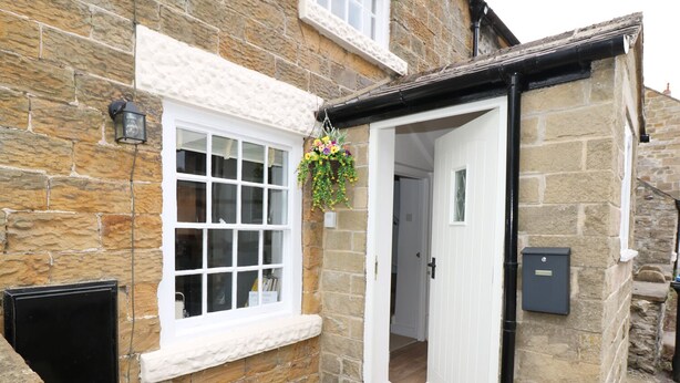 A stone building with a white door, a window with a hanging flower basket, and a black electrical box.
