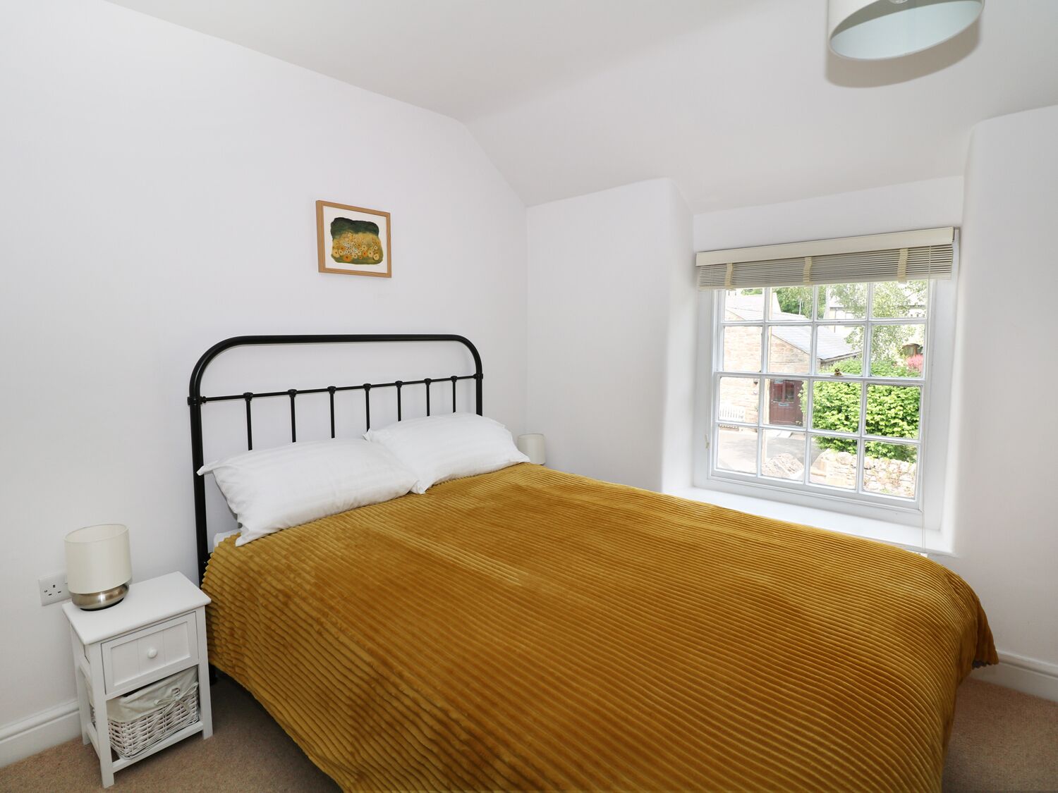 Une chambre avec un lit simple, une table de chevet, une lampe, une fenêtre avec des stores et un tableau au mur.
