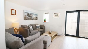 A modern living room with a grey sofa set, a wooden coffee table, and a large window with blinds. - 7 Ocean Gateway (Hythe)