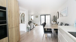 A modern kitchen with wooden cabinets, a dining area with a long table and chairs, and a large window providing natural light. - 7 Ocean Gateway (Hythe)