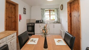 A dining area with a wooden table, two chairs, a vase with flowers, and a kitchen in the background. - The Old Goat Barn at Trout Cottage (Somerton)