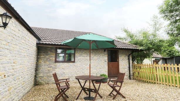 A stone building with a tiled roof, a wooden door, and a window. There is a table and two chairs outside, covered by a green umbrella. - The Old Goat Barn at Trout Cottage (Somerton)