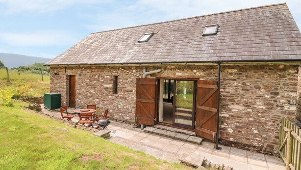 A stone building with a sloped roof, a wooden door, and a small outdoor seating area with a table and chairs. - The Barn (Abergavenny)