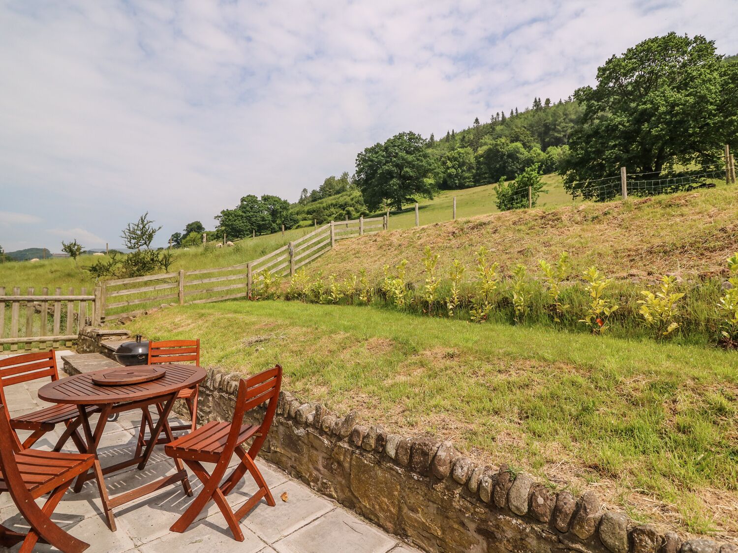 Eine Terrasse mit rundem Tisch und Stühlen, eine Steinmauer und eine sanft abfallende Rasenfläche mit Holzzaun.