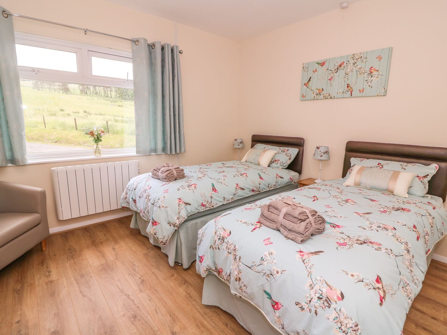 Un dormitorio con dos camas, una ventana con cortinas, un radiador, una silla y un cuadro con motivos florales en la pared.