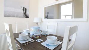 Un coin repas avec une table en bois dressée pour quatre personnes, des chaises blanches, une lampe blanche et une grande fenêtre surmontée d’un miroir.