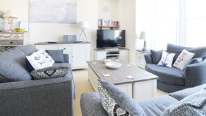 Un salon avec deux canapés gris, une table basse, un téléviseur, une bibliothèque et un lustre.