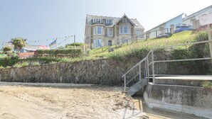Un bâtiment situé sur la côte, doté d’une façade en pierre, d’un escalier menant à un balcon, et d’une plage de sable au premier plan.
