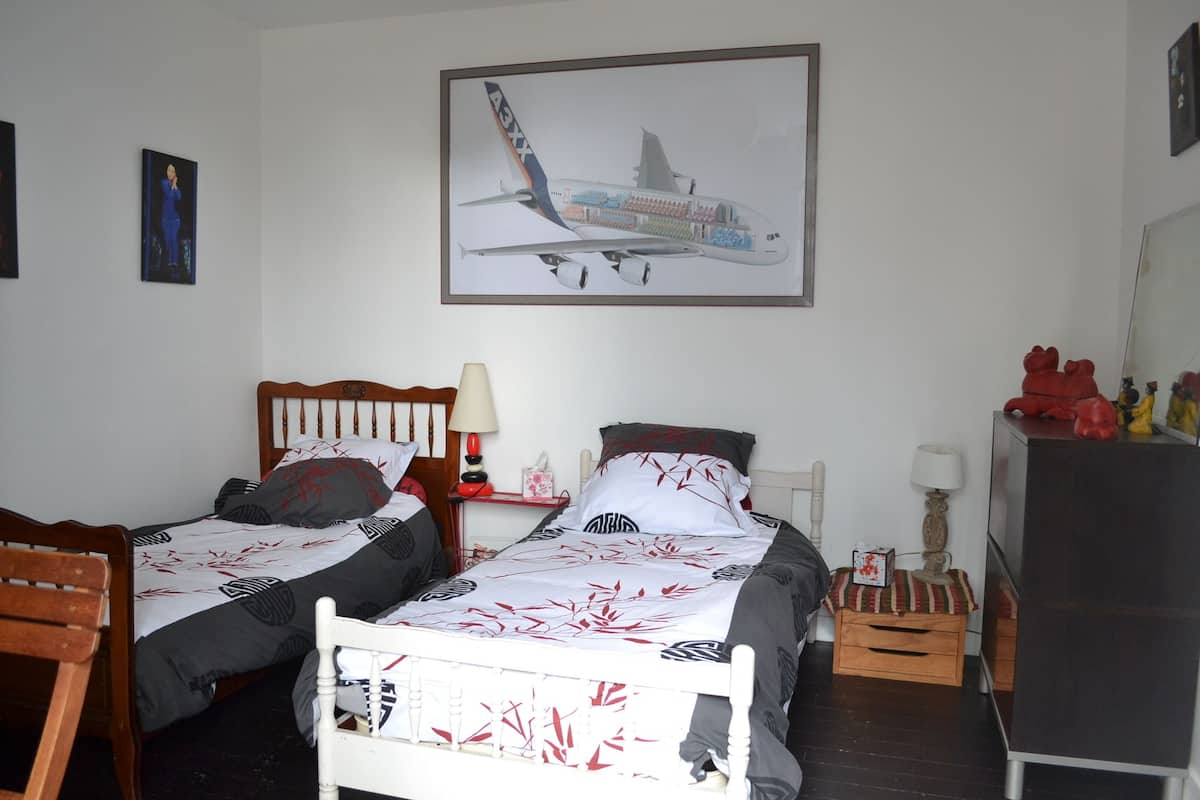 A hotel room with two single beds, a wooden headboard, a nightstand with a lamp, a small table with a lamp, a refrigerator, and a framed airplane picture on the wall.