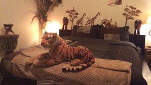 Un tigre recostado en una cama en una habitación con una planta grande, un cuadro de jirafa y una lámpara.