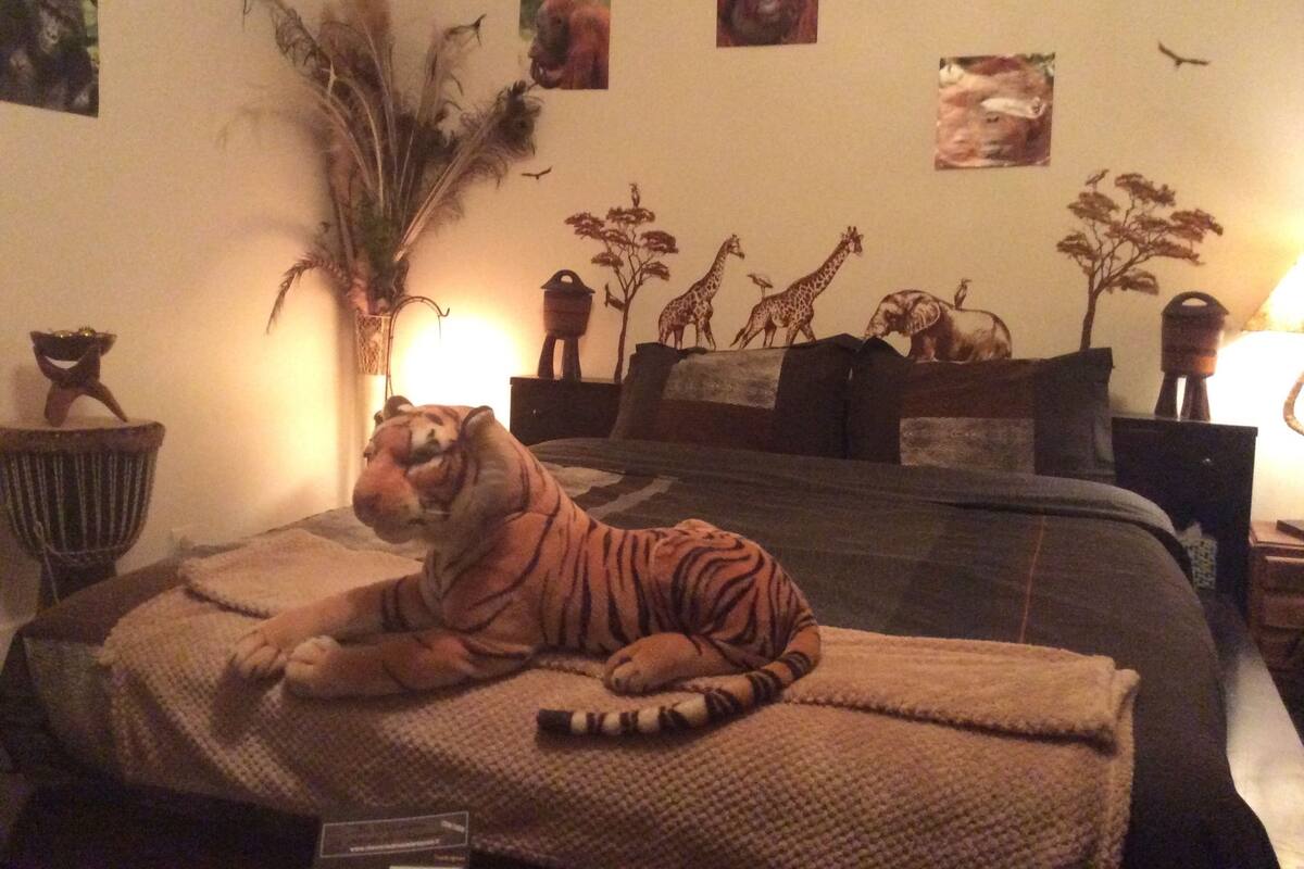 A tiger lying on a bed in a room with a large plant, a giraffe painting, and a lamp.