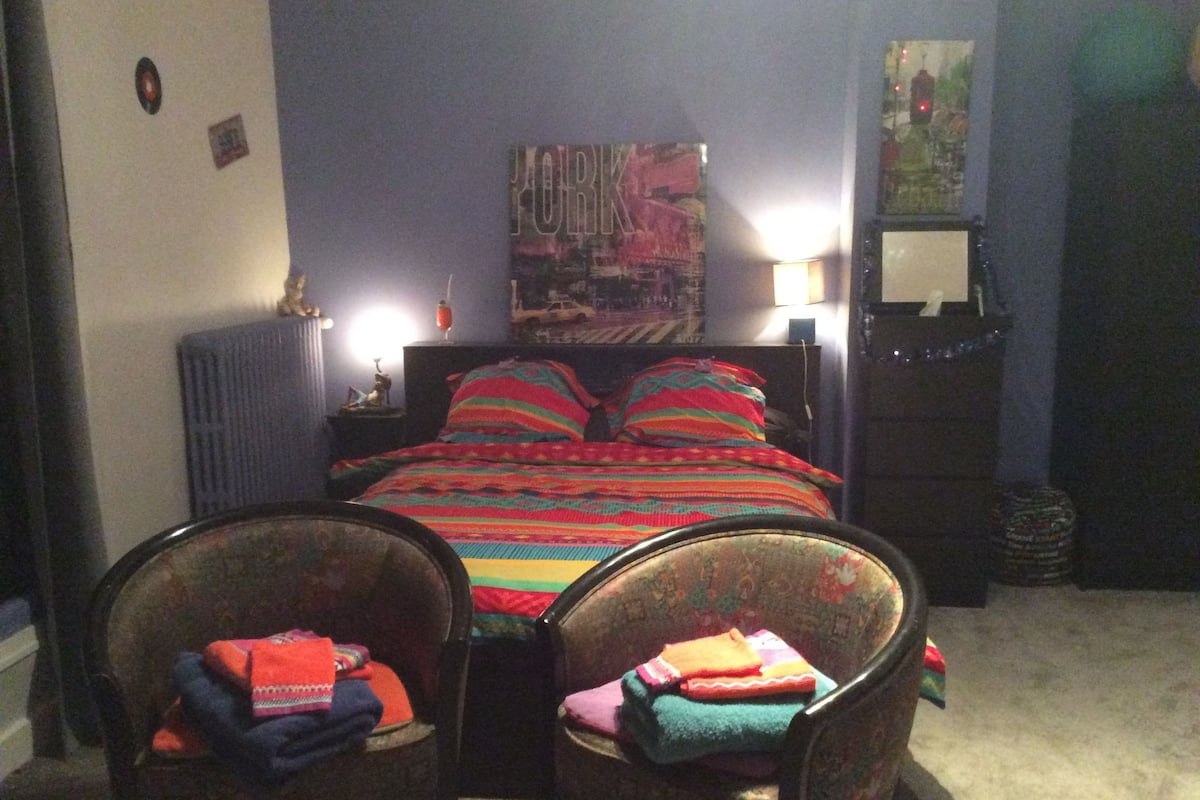 A bedroom with a bed, two chairs, a radiator, a lamp, and a poster on the wall.