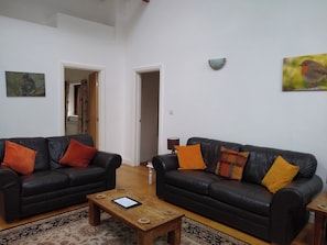 Ein Wohnzimmer mit zwei schwarzen Ledersofas, jeweils mit orangefarbenen und gelben Kissen, einem Holztisch und einem gemusterten Teppich. Zwei Türen führen in andere Räume, eine Wandleuchte und ein gerahmtes Bild sind an der Wand angebracht.