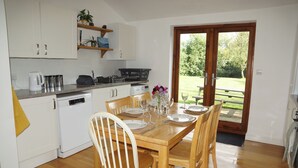 Een eetruimte met een houten tafel voor vier personen, omringd door witte stoelen. Links is een keuken met witte kasten en een aanrecht te zien.