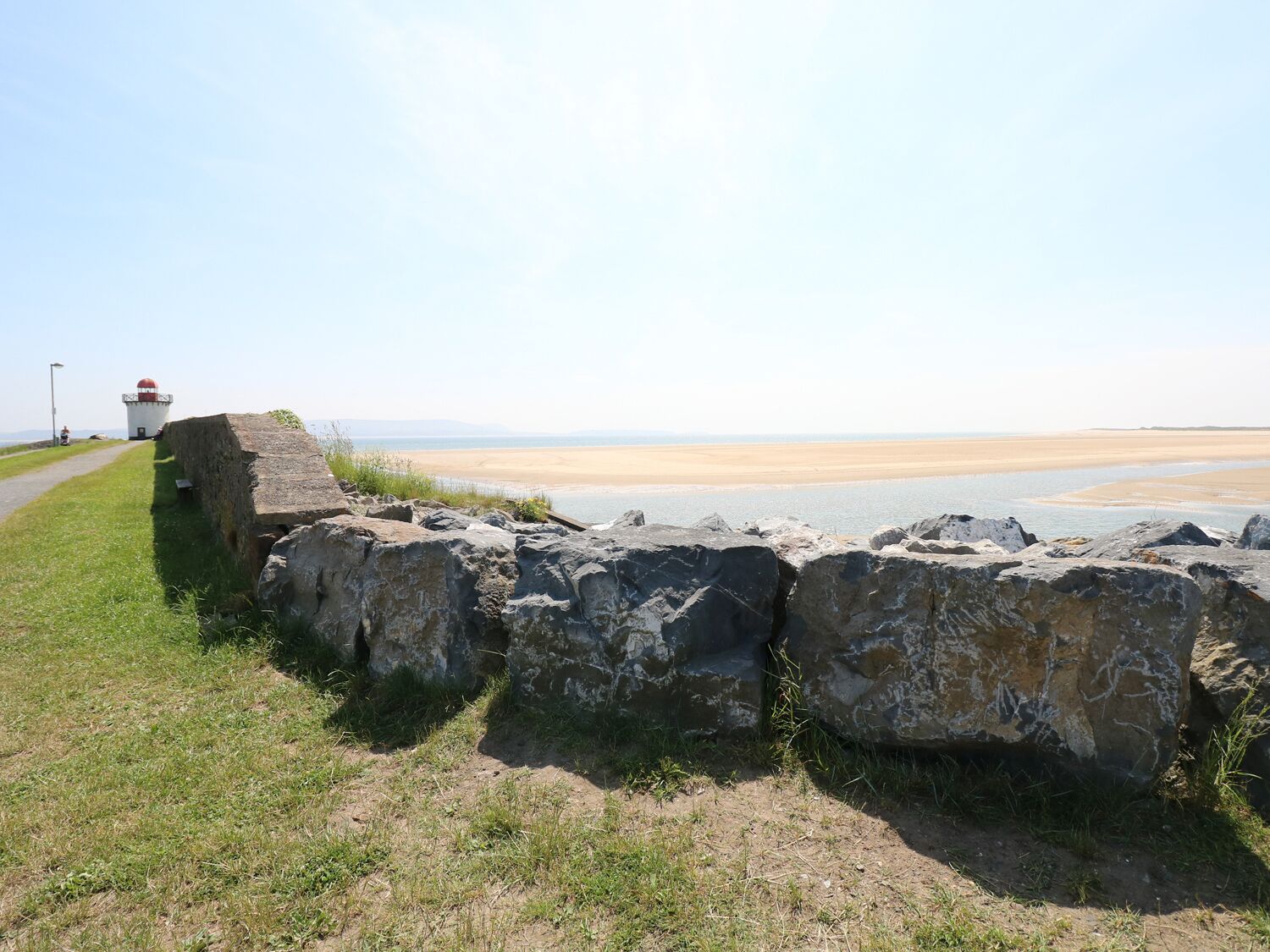 A coastal path with a lighthouse, large rocks, and a sandy beach.