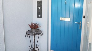 A blue door with the number 5, a small potted plant on a metal table, and a wall-mounted light fixture.