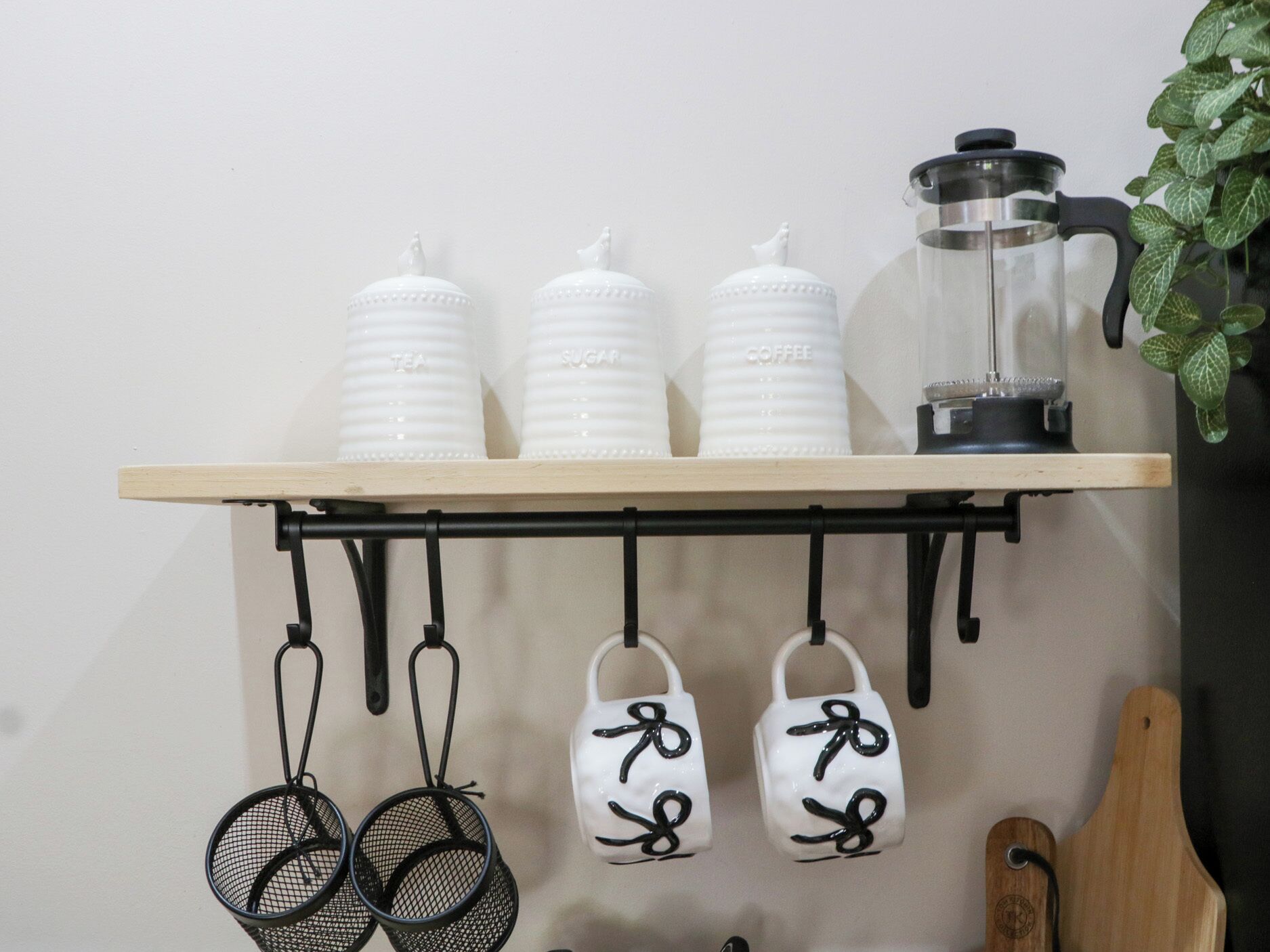 Una repisa de cocina con recipientes blancos etiquetados con té, azúcar y café, un molinillo de café y utensilios de cocina colgantes.