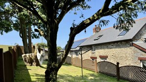 A stone house with a sloped roof, a large tree in the foreground, and a wooden fence. - Caner Bach Lodge (Bridgend)