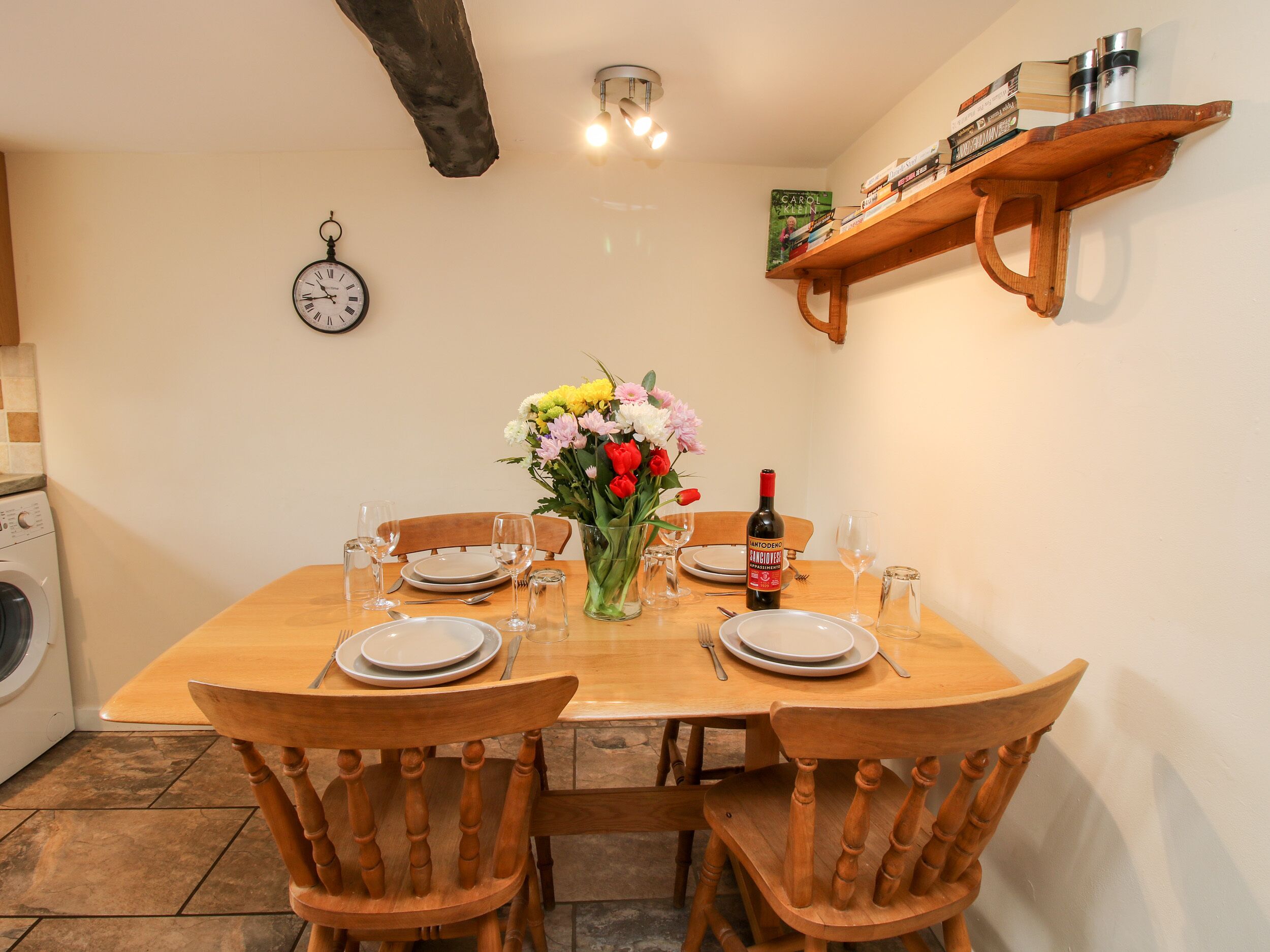 A dining table set for four with chairs, a vase of flowers, wine, and a clock on the wall.