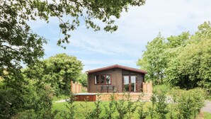 Une petite maison en bois d’un seul étage, dotée d’une terrasse et entourée d’un jardin bien entretenu et d’arbres.